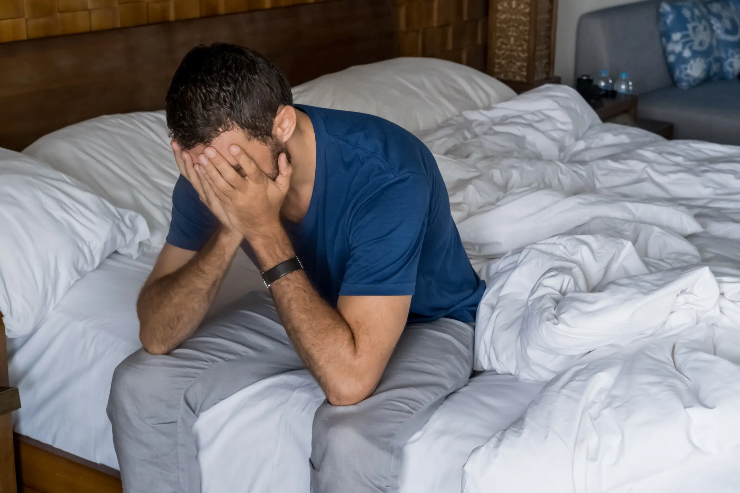 Person sitting on the edge of a bed hands over face overwhelmed
